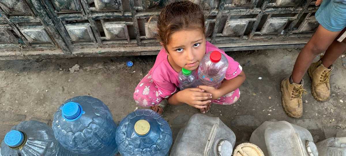La violencia genocida también pasa por la escasez de agua. Una niña espera poder llenar sus botellas vacías. | Foto: UNRWA