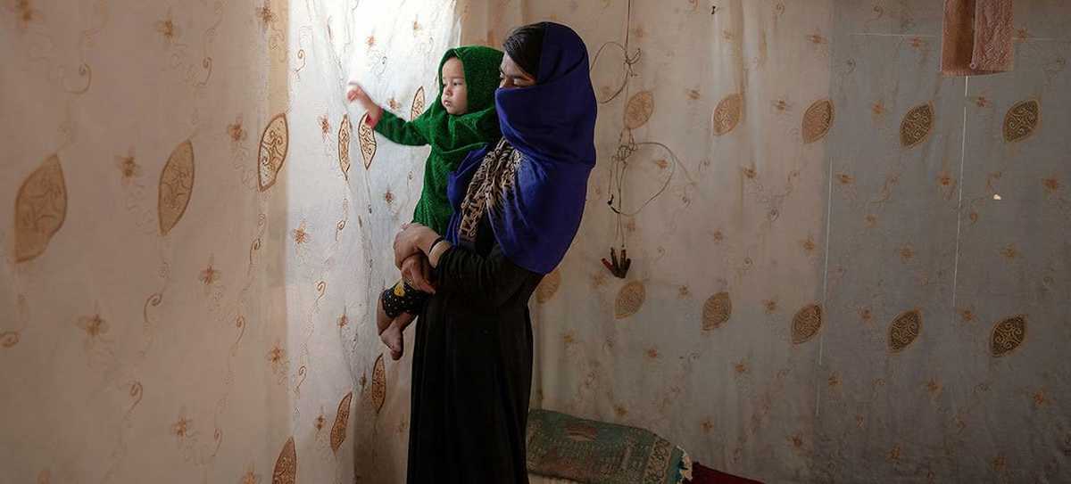 Una madre afgana sostiene a su hija en brazos, mirando la luz desde detrás de su ventana oscurecida. Otra víctima de la "ley de la moralidad". | Foto: UN Women/Sayed Habib Bidell