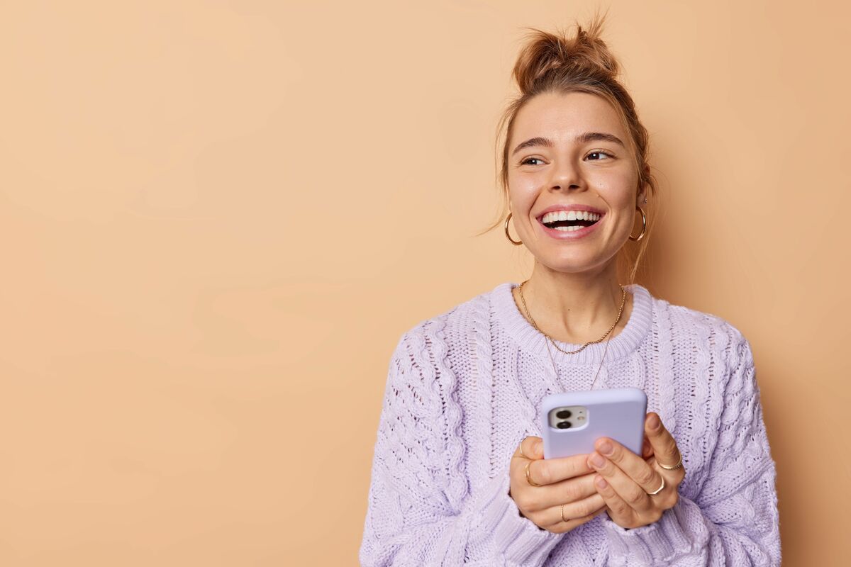 Smiling positive European woman with combed hair uses cellphone enjoys modern technologies holds mobile phone has chat online wears casual sweater isolated over beige background blank space.