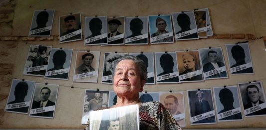 Regina Robledo muestra una fotografía de su padre, Emilio, frente al mural de retratos de las víctimas que preside la entrada a la fosa común donde fueron enterrados. Foto. Jaime Villanueva.